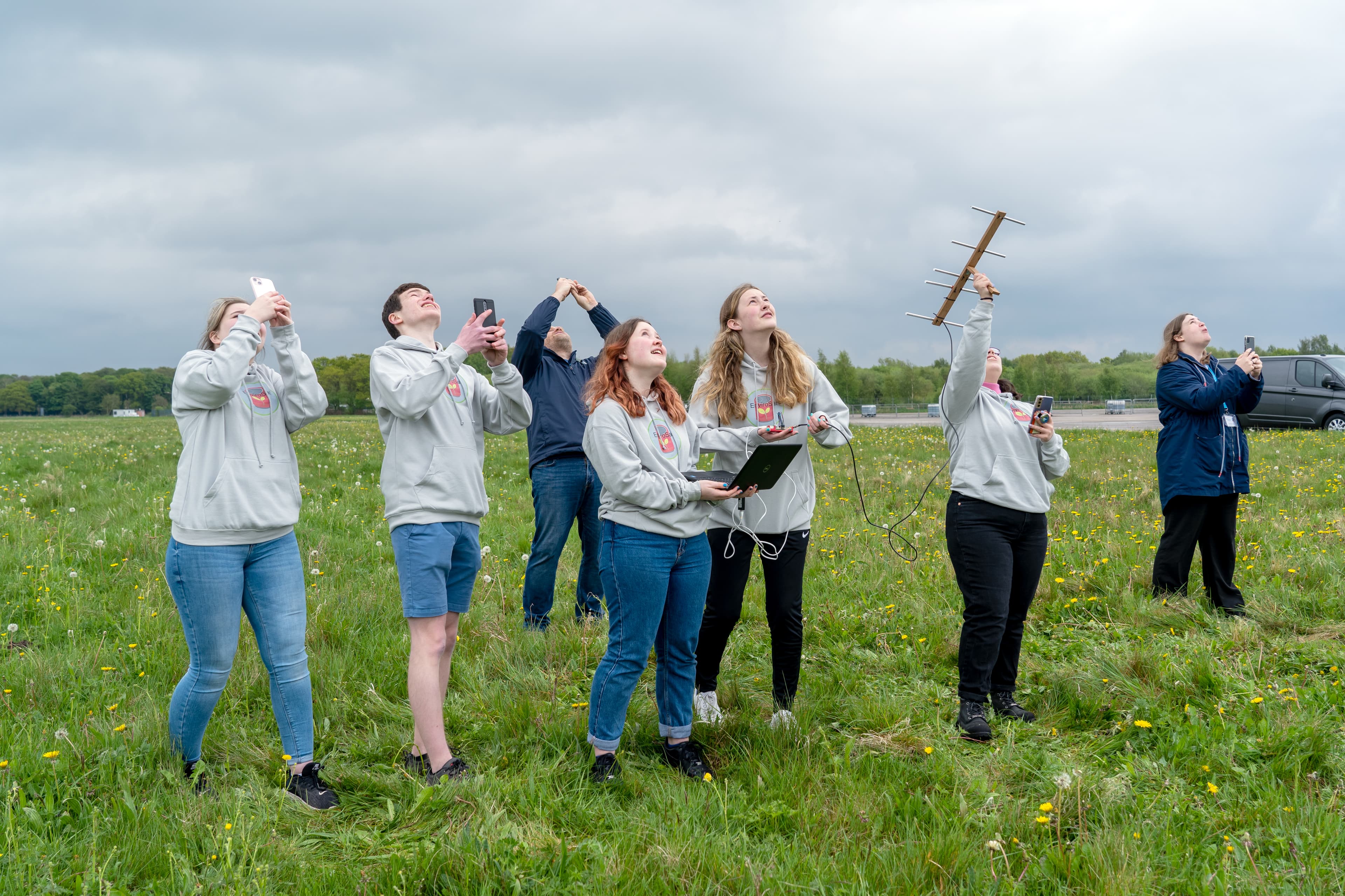 Group of students, looking up at rocket launching in field, one is holding a satellite.